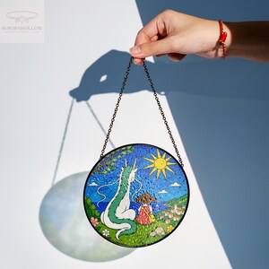 May include: Round stained glass art piece featuring a dragon and a girl looking at a village. The artwork has a black metal chain for hanging. The dragon is white and green, the girl is wearing a red dress, and the sun is yellow.