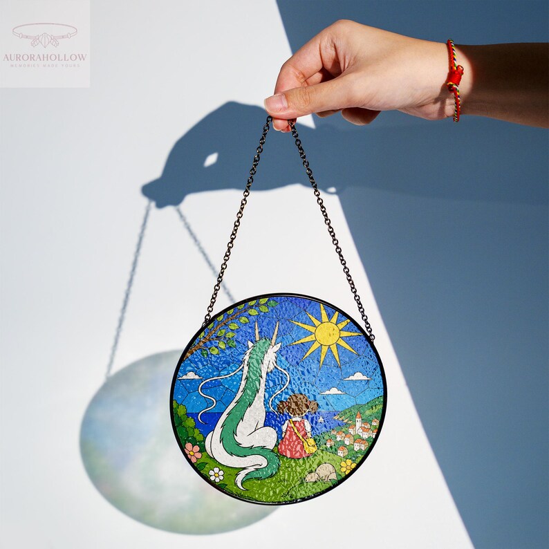 May include: Round stained glass art piece featuring a dragon and a girl looking at a village. The artwork has a black metal chain for hanging. The dragon is white and green, the girl is wearing a red dress, and the sun is yellow.