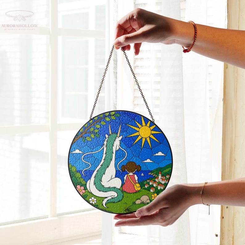 May include: Round stained glass panel featuring a dragon and a girl overlooking a coastal village under a bright sun. The artwork is framed with a black border and suspended by a chain. The color palette includes blue, green, yellow, and pink.