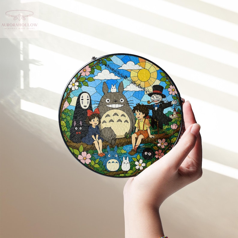May include: Round stained glass art featuring characters from the animated film "My Neighbor Totoro." The artwork depicts Totoro, Mei, Satsuki, and other characters in a colorful, detailed scene with a blue sky and flowers.
