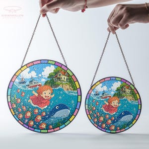 May include: Two round stained glass panels with a chain for hanging. Each panel features a colorful illustration of a girl, a whale, and a coastal scene. The panels have a rainbow-colored border and are held up by hands.