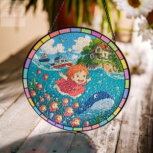 May include: Round stained glass window featuring a vibrant illustration of a girl with red hair, a whale, and boats on a blue ocean. The artwork includes a house on an island, seagulls, and a colorful border. A chain is attached for hanging.