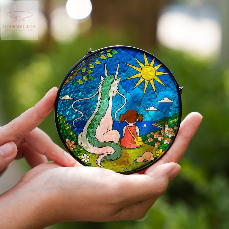 May include: Round stained glass art piece featuring a dragon and a girl overlooking a coastal village. The artwork has a black metal frame and depicts a sunny sky, blue water, and green hills. The piece is held in open hands.