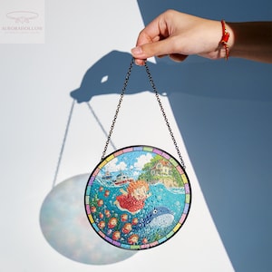 May include: Round stained glass art piece featuring a whimsical underwater scene with a girl, whale, and other characters. The artwork is held by a chain and has a colorful border. The image is set against a white and blue background.