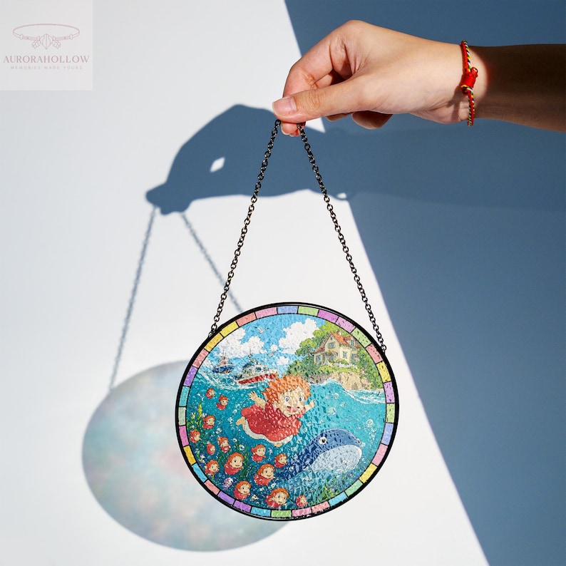 May include: Round stained glass art piece featuring a whimsical underwater scene with a girl, whale, and other characters. The artwork is held by a chain and has a colorful border. The image is set against a white and blue background.