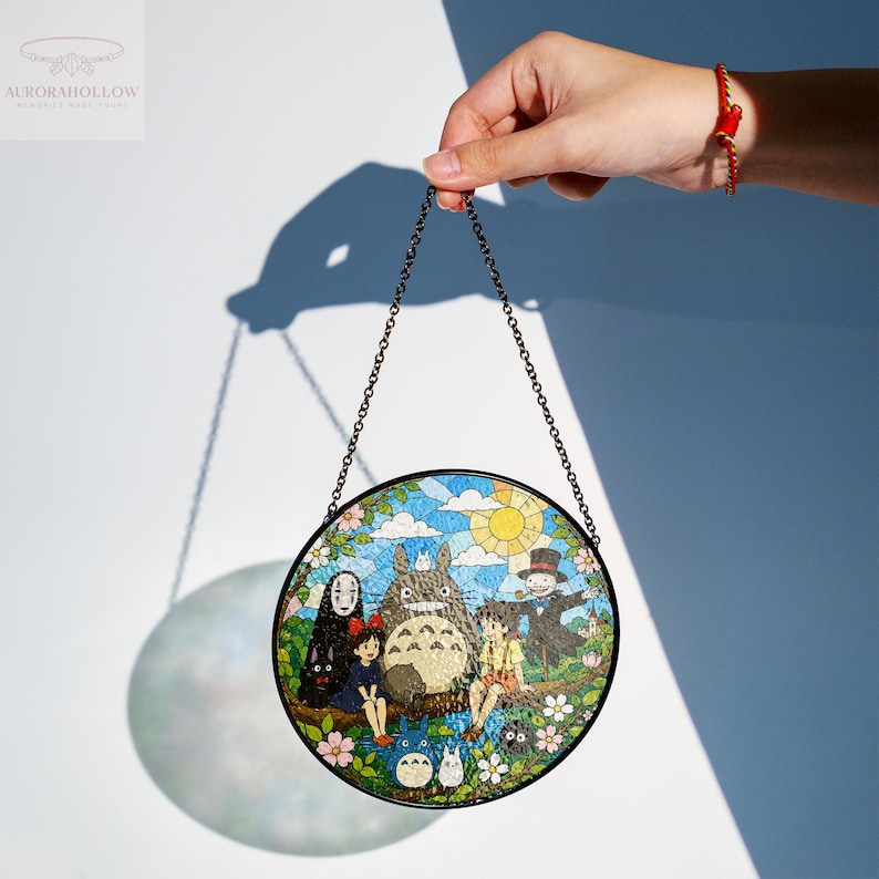 May include: A round stained glass panel featuring characters from the animated film "My Neighbor Totoro." The artwork depicts Totoro, Mei, Satsuki, and other characters in a colorful mosaic design. The panel is held by a chain.