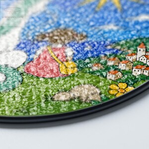 May include: A colorful, mosaic-style artwork depicting a whimsical landscape. The image features a figure in a red dress, a yellow broom, and a village with white buildings and orange roofs. The sky is blue with a yellow sun. The artwork is framed with a black border.
