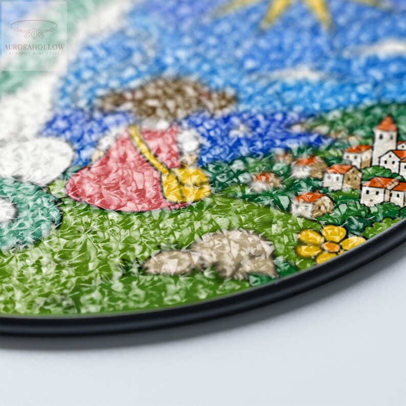 May include: A colorful, mosaic-style artwork depicting a whimsical landscape. The image features a figure in a red dress, a yellow broom, and a village with white buildings and orange roofs. The sky is blue with a yellow sun. The artwork is framed with a black border.