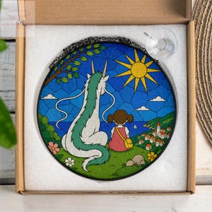 May include: Round stained glass art piece featuring a dragon and a girl overlooking a coastal town. The artwork has a blue sky with a yellow sun, white clouds, and green hills. The piece is in a cardboard box.