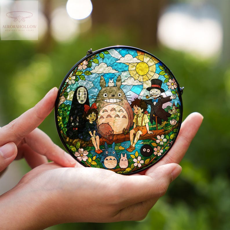 May include: A round stained glass panel featuring characters from the animated film "My Neighbor Totoro." The artwork depicts Totoro, Mei, Satsuki, and other characters in a colorful, detailed scene with a sunny sky and floral accents. The panel is held in a person's hands.