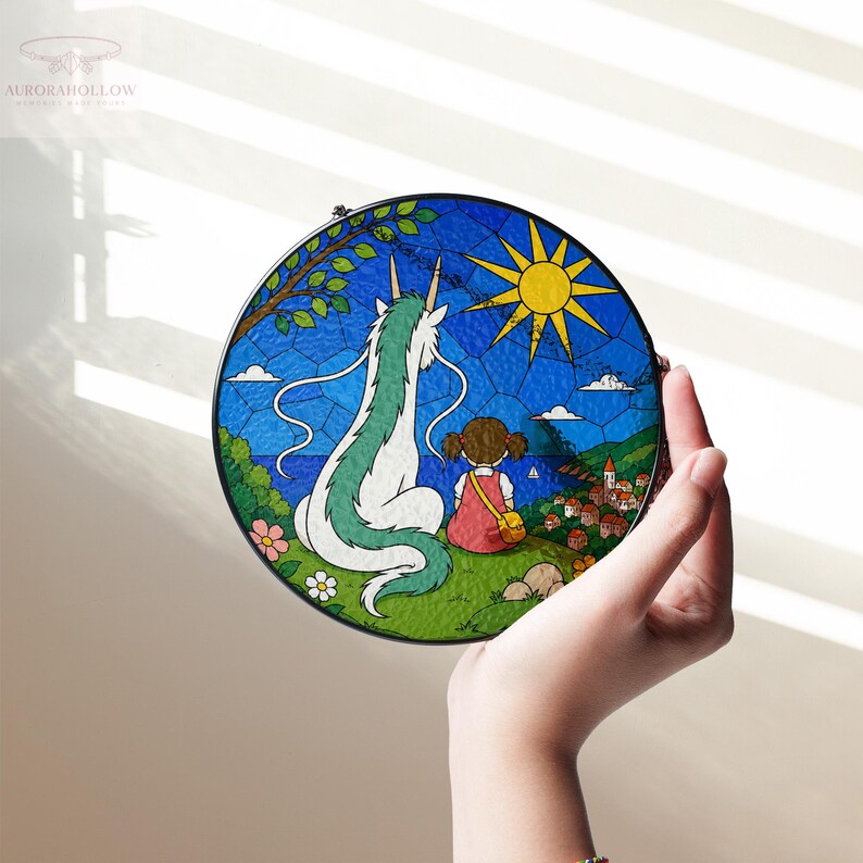 May include: A round stained glass panel featuring a scene from a popular animated film. The artwork depicts a girl and a dragon-like creature overlooking a coastal town under a bright sun. The panel is held by a hand.