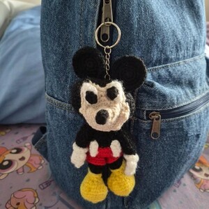 May include: A handmade crochet Mickey Mouse keychain. The keychain features a black head and ears, a beige face, white gloves, red shorts, and yellow shoes. It is attached to a silver keyring and hangs from a denim backpack.