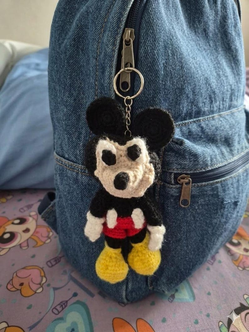 May include: A handmade crochet Mickey Mouse keychain. The keychain features a black head and ears, a beige face, white gloves, red shorts, and yellow shoes. It is attached to a silver keyring and hangs from a denim backpack.
