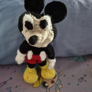May include: A handmade crochet Mickey Mouse doll. The doll features black ears, a black head, and a tan face. It wears red shorts, yellow shoes, and white gloves. The doll is standing on a patterned surface.