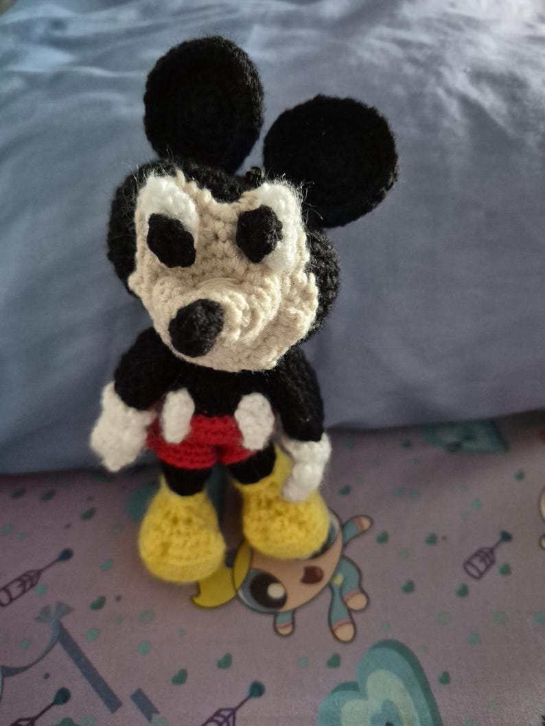 May include: A handmade crochet Mickey Mouse doll. The doll features black ears, a black head, and a tan face. It wears red shorts, yellow shoes, and white gloves. The doll is standing on a patterned surface.