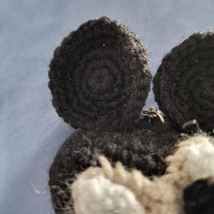 May include: Close-up of a handmade crochet animal head with black and white details. The ears are black and circular, with a spiral pattern. The face has white and tan sections, with black accents.