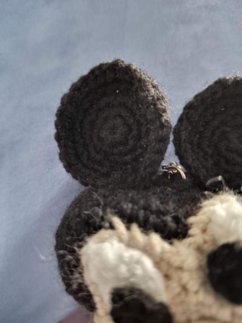 May include: Close-up of a handmade crochet animal head with black and white details. The ears are black and circular, with a spiral pattern. The face has white and tan sections, with black accents.