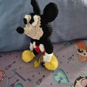 May include: A handmade crochet Mickey Mouse figure. The figure features black, white, red, and yellow yarn. The character has large black ears, a white face, and yellow shoes. The figure is standing on a patterned surface.