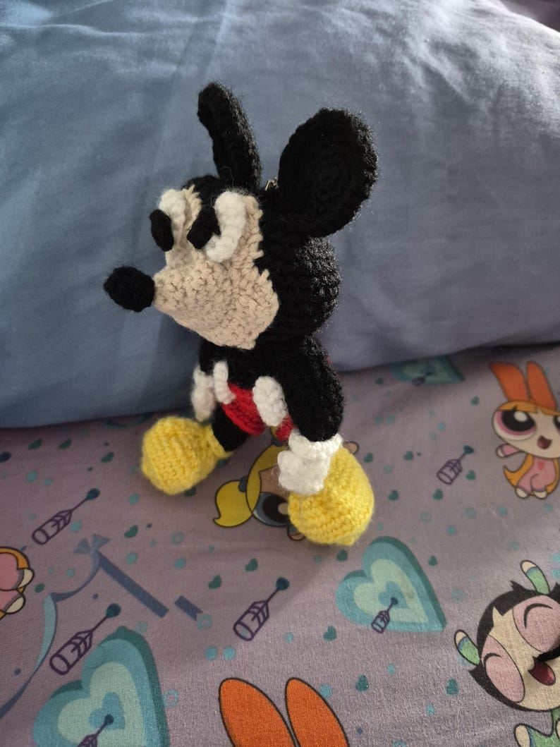 May include: A handmade crochet Mickey Mouse figure. The figure features black, white, red, and yellow yarn. The character has large black ears, a white face, and yellow shoes. The figure is standing on a patterned surface.