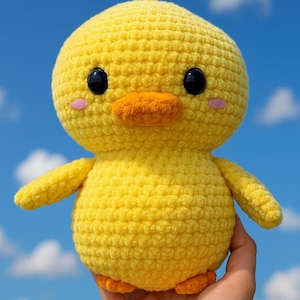 May include: A handmade, yellow crochet duckling with black eyes, an orange beak, and small pink cheeks. The duckling has small wings and feet. The background is a bright blue sky with fluffy white clouds.