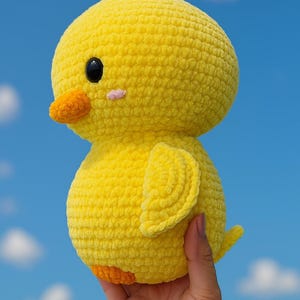 May include: A handmade, yellow crochet chick with an orange beak and feet, and a small pink cheek. The chick has a round body and head, and small wings. The background is a bright blue sky with white clouds.