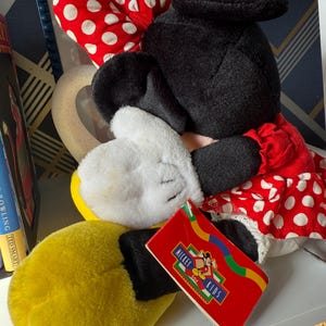 May include: A plush Minnie Mouse doll with a red and white polka dot bow, red dress with white polka dots, and yellow gloves. The doll has a black head and ears, and a tag that says "Mickey Toy Kids".