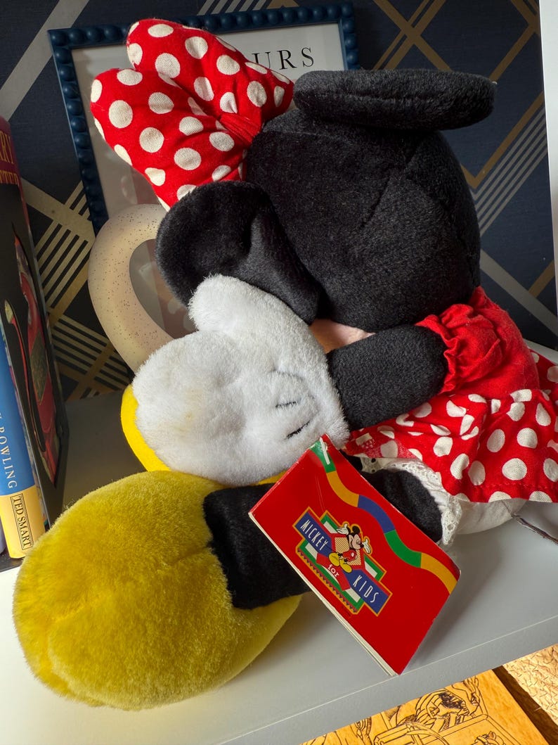 May include: A plush Minnie Mouse doll with a red and white polka dot bow, red dress with white polka dots, and yellow gloves. The doll has a black head and ears, and a tag that says "Mickey Toy Kids".