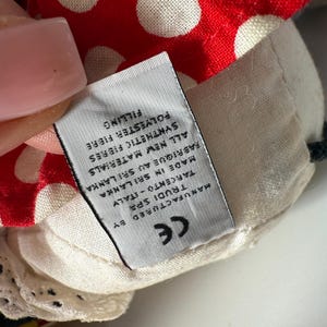 May include: Close-up of a plush toy's head, featuring a red fabric with white polka dots. The toy has a cream-colored fabric with a tag that reads "MANUFACTURED BY TRUDI SPA TARCENTO-ITALY MADE IN SRI LANKA".