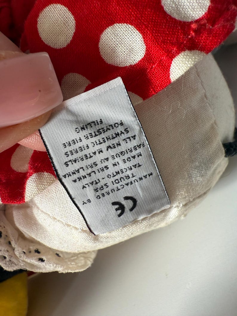 May include: Close-up of a plush toy's head, featuring a red fabric with white polka dots. The toy has a cream-colored fabric with a tag that reads "MANUFACTURED BY TRUDI SPA TARCENTO-ITALY MADE IN SRI LANKA".