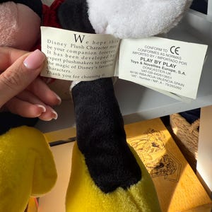 May include: A plush Disney character with a black head, white face, and yellow body. The character has a red bow tie and a tag with text. The tag reads "Disney Plush Character may be your companion forever."