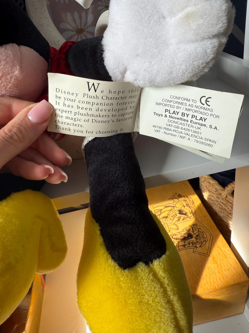 May include: A plush Disney character with a black head, white face, and yellow body. The character has a red bow tie and a tag with text. The tag reads "Disney Plush Character may be your companion forever."