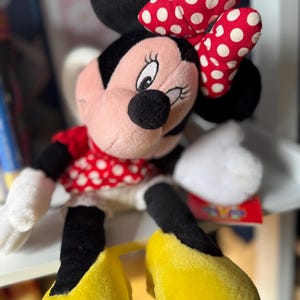May include: A plush Minnie Mouse doll with a red and white polka dot bow, dress, and matching shoes. The doll has black ears, a black nose, and white gloves. The doll is sitting on a shelf.