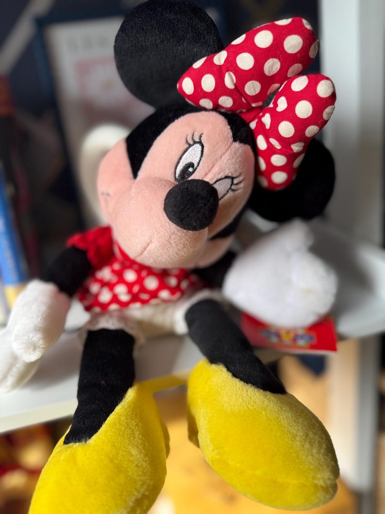 May include: A plush Minnie Mouse doll with a red and white polka dot bow, dress, and matching shoes. The doll has black ears, a black nose, and white gloves. The doll is sitting on a shelf.