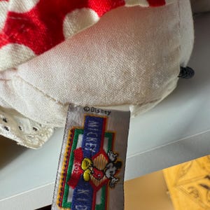May include: Close-up of a plush toy featuring a red and white polka-dotted pattern. The toy has a tag with the Disney logo, the words "Mickey Kids", and an image of Mickey Mouse.