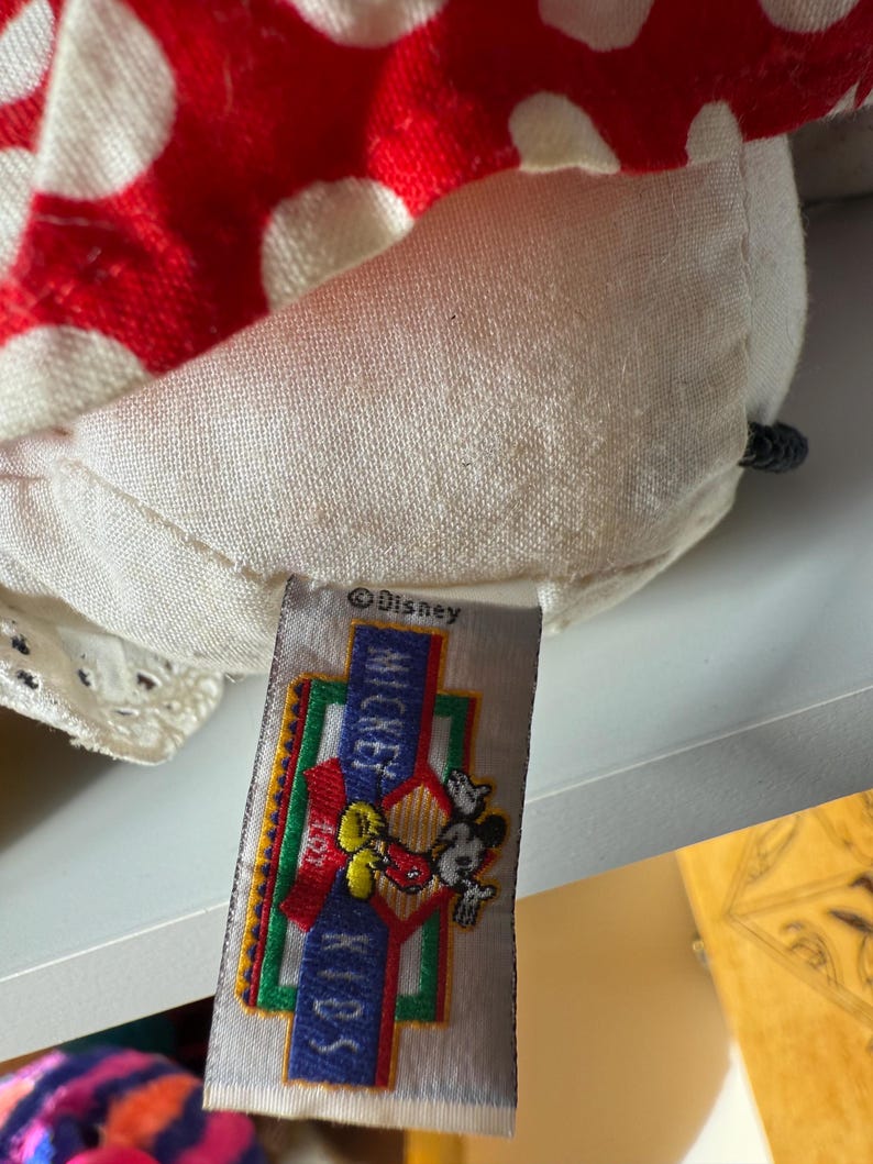 May include: Close-up of a plush toy featuring a red and white polka-dotted pattern. The toy has a tag with the Disney logo, the words "Mickey Kids", and an image of Mickey Mouse.