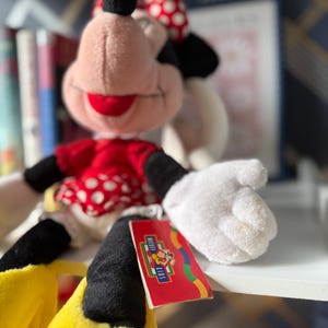 May include: A Minnie Mouse plush doll with a red and white polka dot dress and bow. The doll has black ears, arms, and a tail, with yellow shoes and white gloves. A red tag with the Mickey Kids logo is attached.