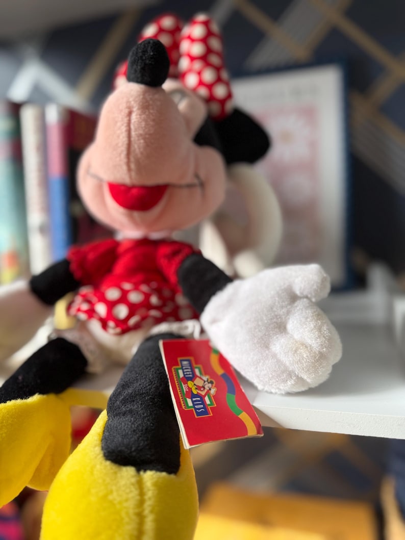 May include: A Minnie Mouse plush doll with a red and white polka dot dress and bow. The doll has black ears, arms, and a tail, with yellow shoes and white gloves. A red tag with the Mickey Kids logo is attached.