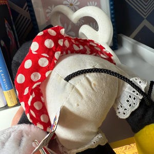 May include: A Minnie Mouse plush doll, featuring a red polka dot bow, white dress with lace trim, and black gloves. The doll has a tag attached.