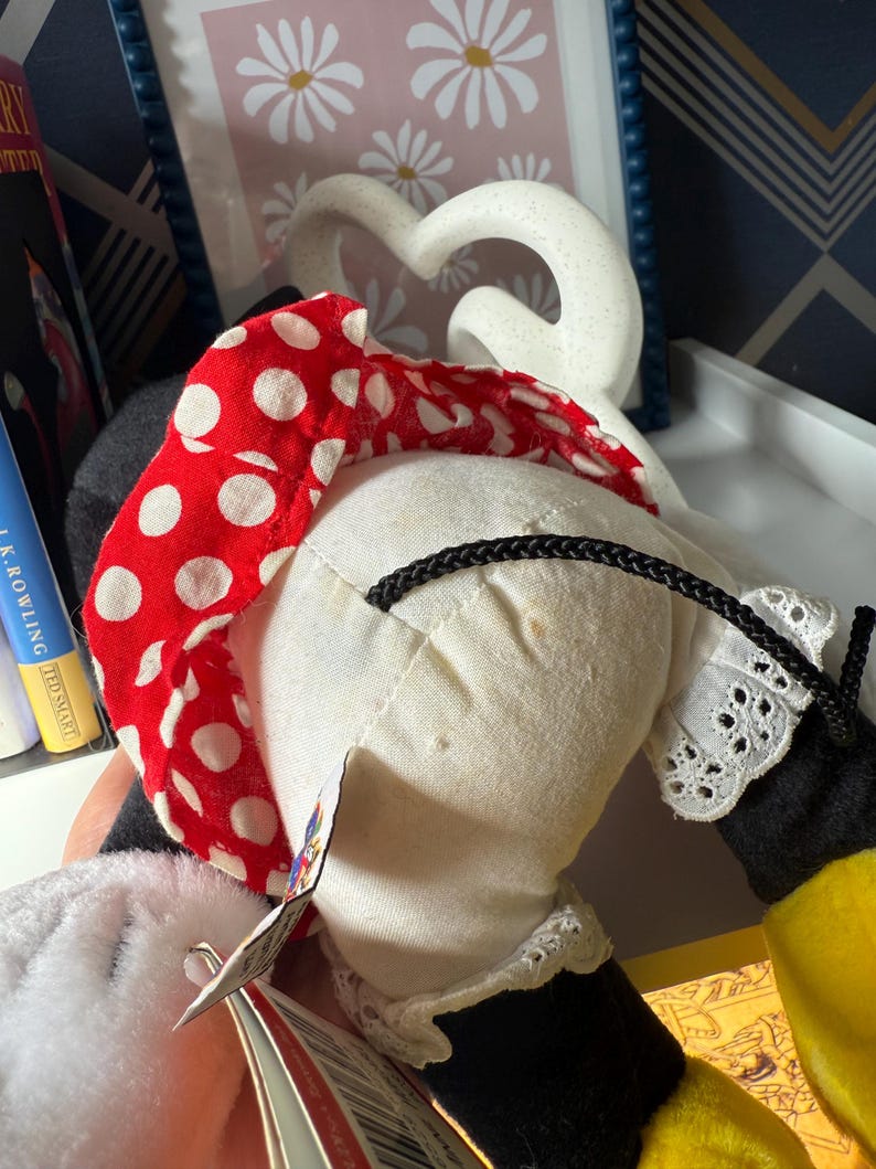 May include: A Minnie Mouse plush doll, featuring a red polka dot bow, white dress with lace trim, and black gloves. The doll has a tag attached.