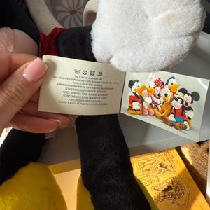 May include: A plush Mickey Mouse doll with black, white, red, and yellow features. The doll has a white glove, black legs, and yellow feet. A tag with text and a picture of Disney characters is attached.