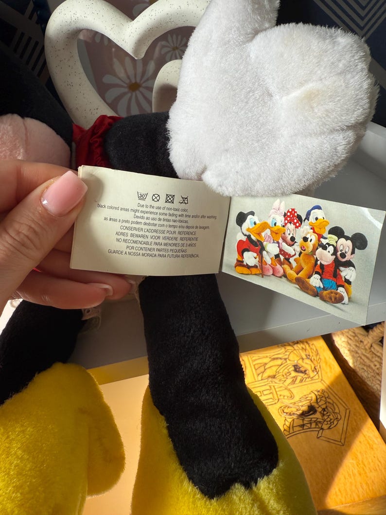 May include: A plush Mickey Mouse doll with black, white, red, and yellow features. The doll has a white glove, black legs, and yellow feet. A tag with text and a picture of Disney characters is attached.