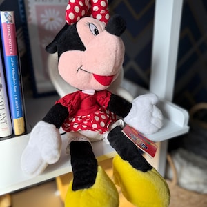 May include: A plush Minnie Mouse doll, featuring a red dress with white polka dots, a matching bow, and yellow shoes. The doll has black ears, a pink face, and white gloves. A tag is attached to the doll's leg.