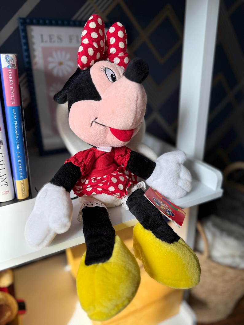 May include: A plush Minnie Mouse doll, featuring a red dress with white polka dots, a matching bow, and yellow shoes. The doll has black ears, a pink face, and white gloves. A tag is attached to the doll's leg.