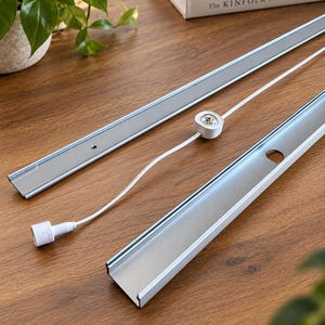 May include: Two long, narrow, silver-coloured metal channels with white edges, accompanied by a small white circular component connected to a white electrical cord and a white plug, designed for lighting installation or shelving support systems.