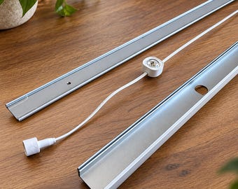 May include: Two long, narrow, silver-coloured metal channels with white edges, accompanied by a small white circular component connected to a white electrical cord and a white plug, designed for lighting installation or shelving support systems.