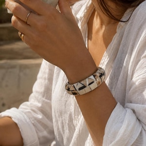 May include: A thick bangle bracelet featuring a geometric mosaic pattern of inlaid cream, tan, and black segments. The bracelet has a smooth, rounded profile and a polished finish, showcasing a repeating triangular and linear design across its surface.