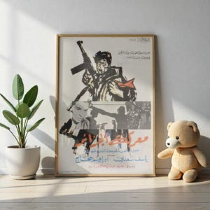 May include: A framed vintage poster with Arabic text and illustrations. The artwork features a man holding a rifle, with other figures and text below. The poster is in a wooden frame and leans against a white wall.