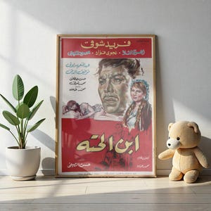 Puede incluir: Un póster de película vintage enmarcado con texto e ilustraciones árabes. El póster presenta un retrato de hombre, una mujer y una figura reclinada, sobre un fondo rojo. Un oso de peluche y una planta en maceta están en primer plano.