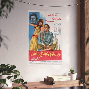 Peut inclure: Affiche de film vintage avec texte arabe et illustration colorée de trois personnages. Le titre "Rage Over a Hot Plate" est en rouge. L'affiche est accrochée à un mur blanc, avec une étagère en bois et des plantes devant, pour une ambiance rétro.