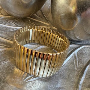 May include: A gold-coloured, expandable bracelet with a ribbed design. The bracelet is made of rectangular links that expand to fit the wrist. The jewellery is displayed on a silver, textured surface.